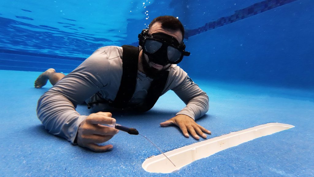 A person underwater in a swimming pool wears a mask and holds a tool while inspecting a pool component.