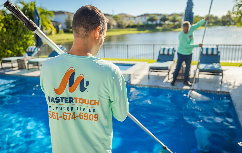 Two men cleaning a pool with nets and poles, wearing light green shirts with a company logo and phone number. They are in a backyard with a lake view in the background.