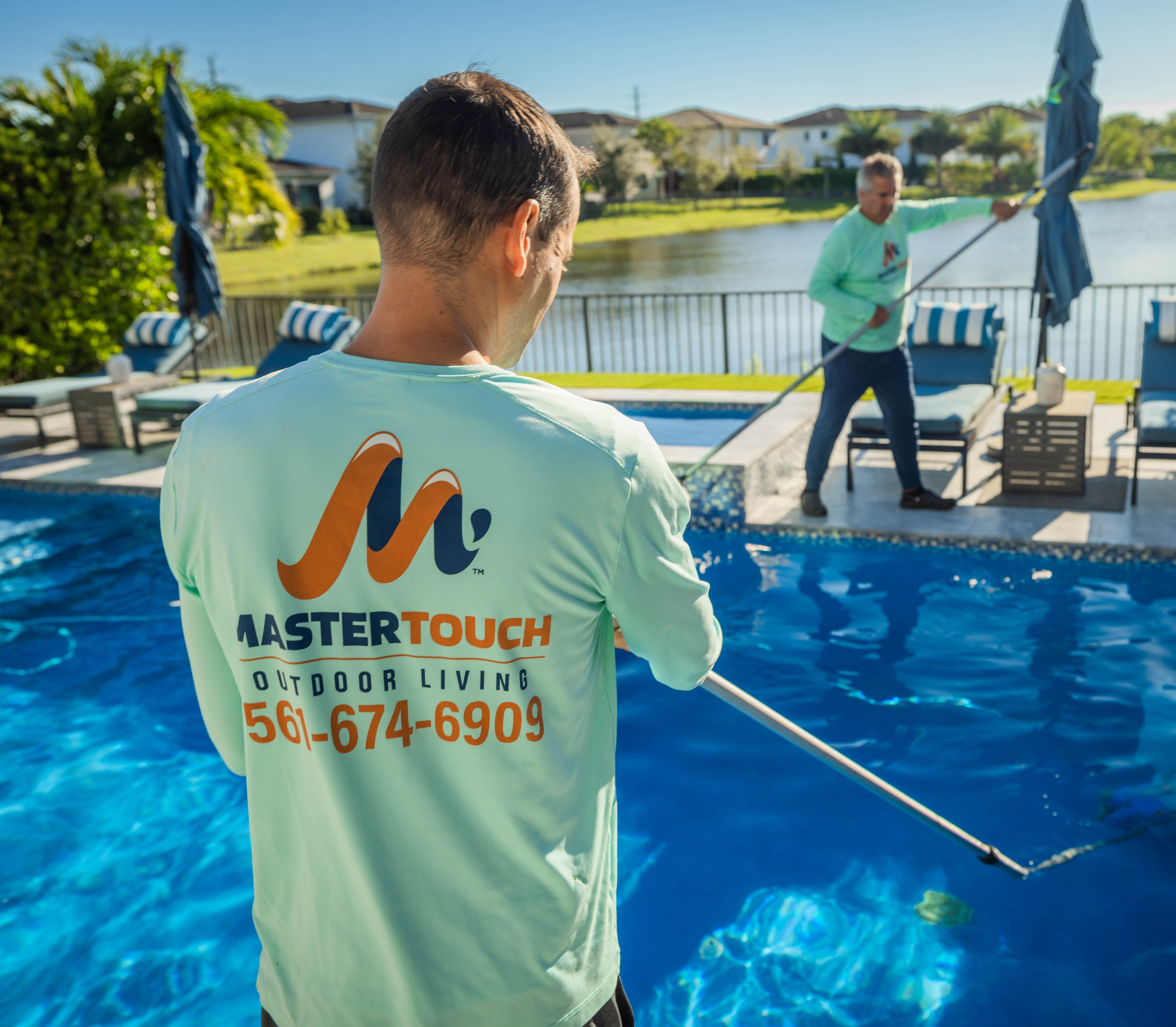 wo people cleaning a pool in a backyard, one wearing a Master Touch Outdoor Living shirt with a phone number. The pool overlooks a scenic lake with palm trees and houses in the background, and there are lounge chairs with umbrellas by the poolside.